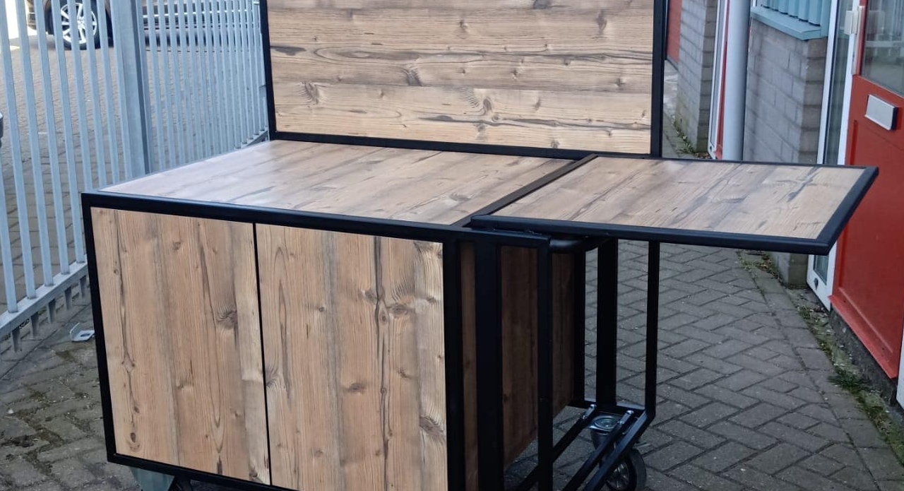 Verrijdbare koffiekar in houtlook, afgewerkt met Plastica HPL 0921SU. Voorzien van opklapbaar werkblad en zwart metalen frame.